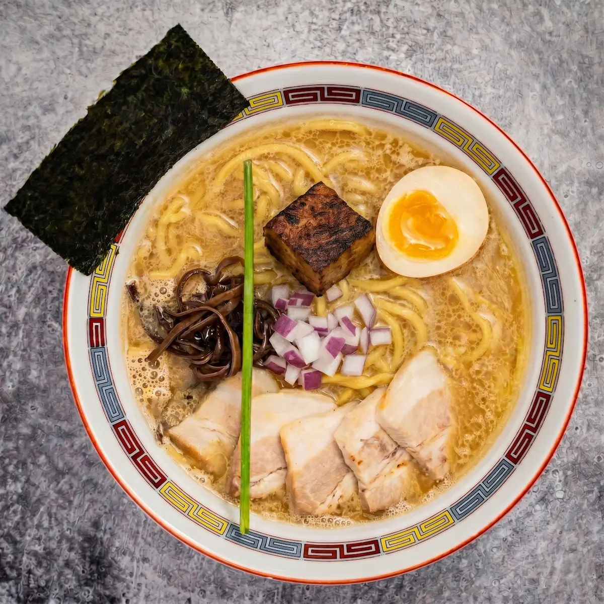 Tonkotsu Shoyu - A bowl of rich pork and soy sauce broth ramen with thick noodles, pork chashu, diced red onion, chive, kikurage mushroom, nori, and a soft-boiled egg