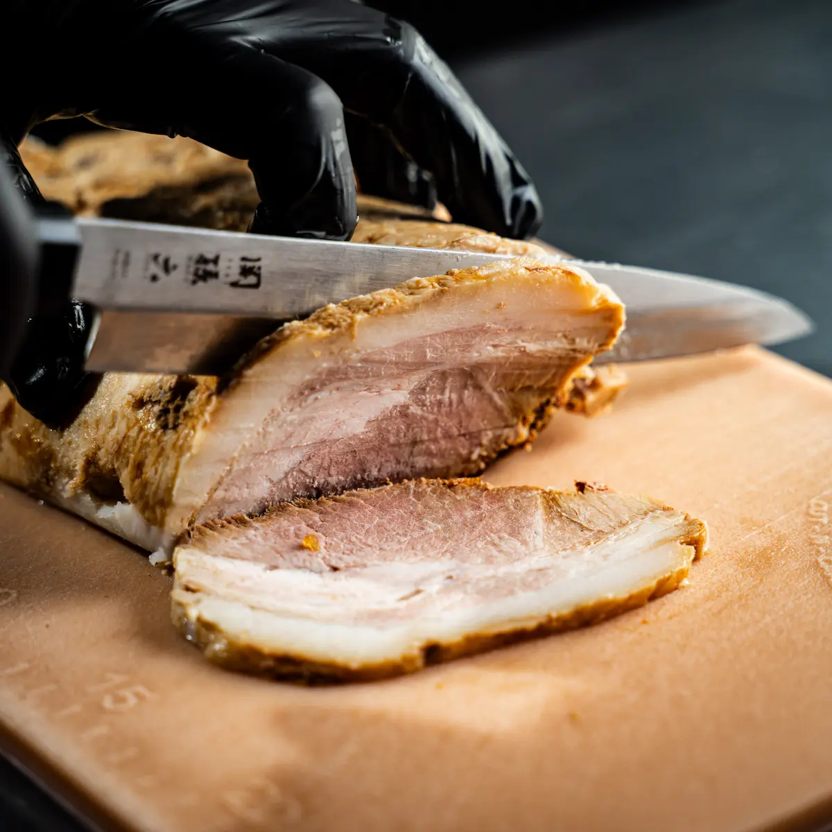 A close-up of a chef wearing black gloves slicing a thick, juicy pork chashu block with a Japanese knife at Japanese Ramen Gachi. Sliced to order to prevent oxidation and ensure peak flavor.